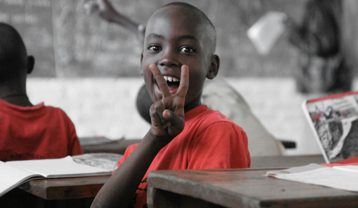 Jeune garçon en uniforme rouge faisant le signe de la paix dans une salle de classe en Afrique — accès à l’éducation solidaire.